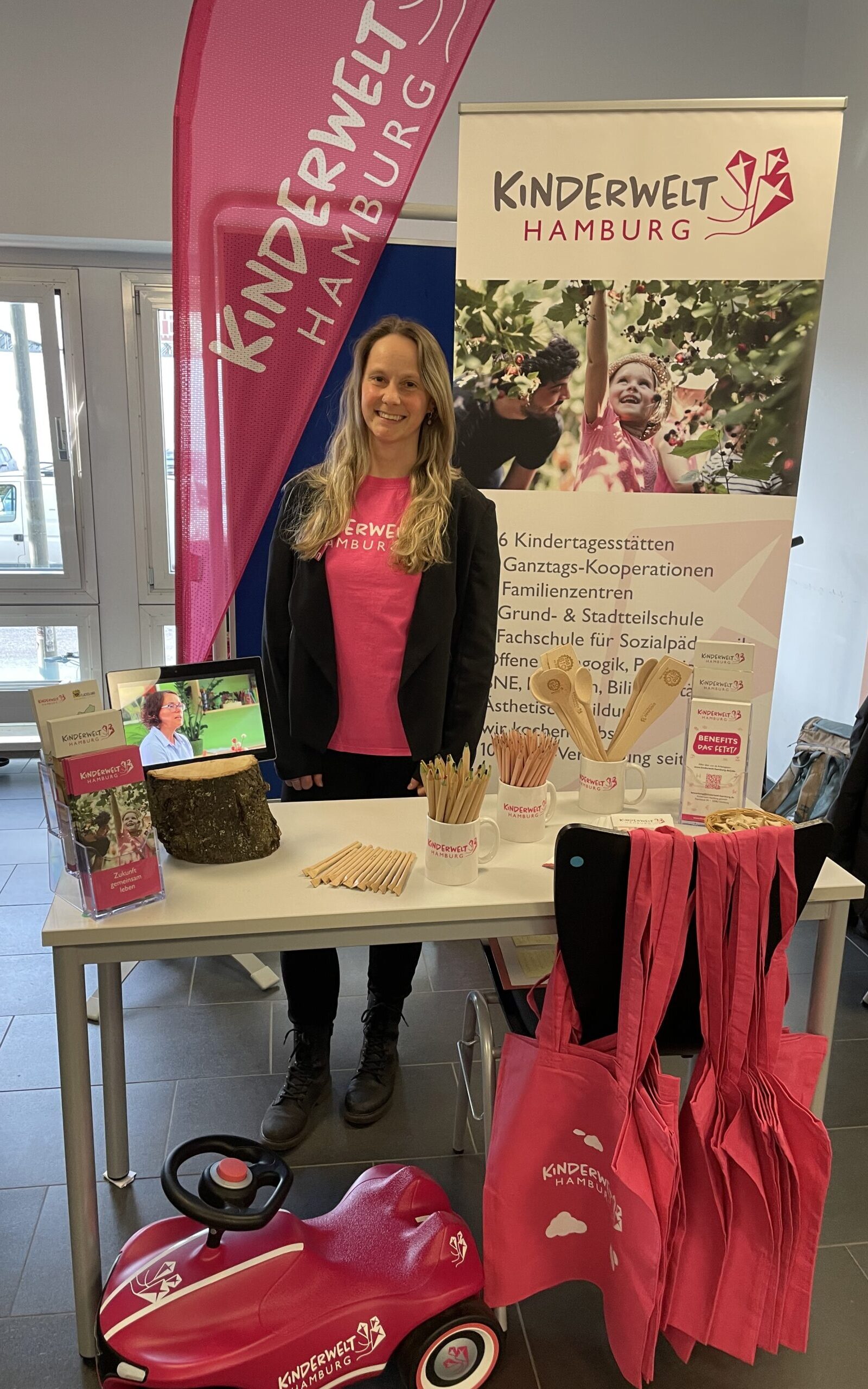 Messestand von Kinderwelt Hamburg: Person im pinken T‑Shirt hinter einem Stand mit Flyern, Holzstiften und pinken Stofftaschen; daneben Banner und pinkes Bobbycar