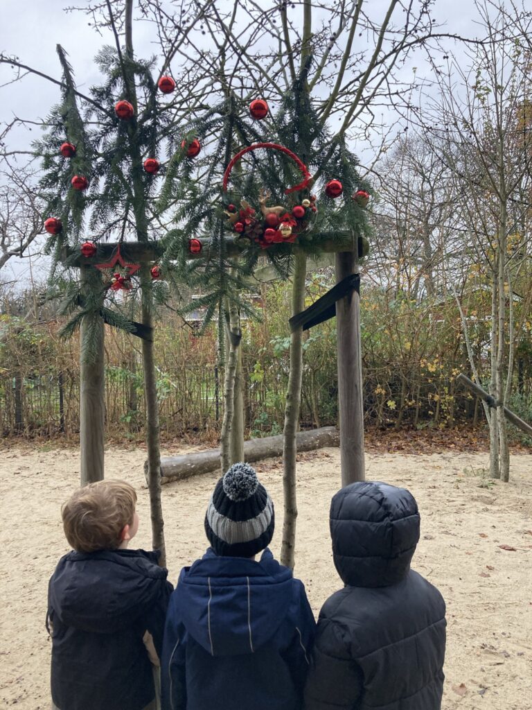 Drei Kinder stehen im Garten vor einer mit Tannenzweigen und roten Weihnachtskugeln geschmückten Holzstruktur und betrachten den weihnachtlichen Schmuck.