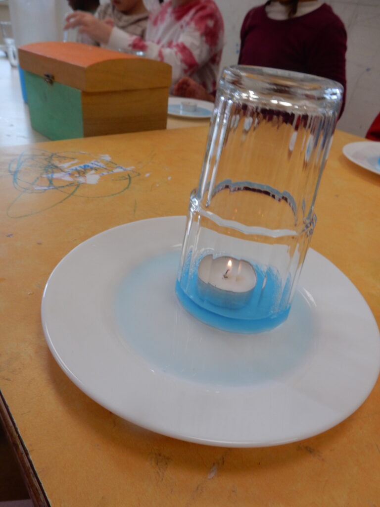 Ein Teelicht schwimmt in gefärbtem Wasser auf einem weißen Teller. Ein umgestülptes Glas steht über der Kerze und umschließt sie. Im Hintergrund sitzen unscharf zwei Kinder an einem Tisch.