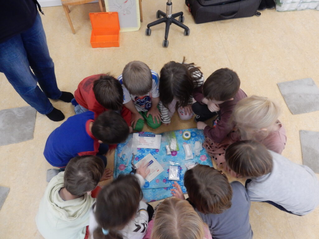 Eine Gruppe von Kindern sitzt im Kreis auf dem Boden einer Kita und betrachtet gemeinsam verschiedene Erste‑Hilfe‑Materialien, die in der Mitte auf einer Decke liegen. Ein erwachsener Mensch steht am Rand des Kreise