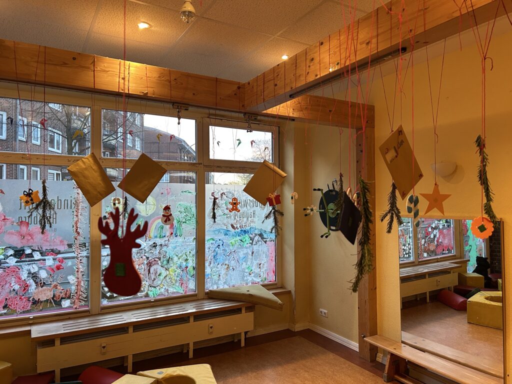 Bewegungsraum der Kita mit einem besonderen Adventskalender: Umschläge, Anhänger und Tannenzweige hängen von der Decke, vor großen Fenstern mit bunten Kinder-Malereien.