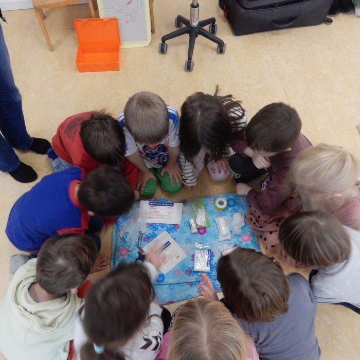 Eine Gruppe von Kindern sitzt im Kreis auf dem Boden einer Kita und betrachtet gemeinsam verschiedene Erste‑Hilfe‑Materialien, die in der Mitte auf einer Decke liegen. Ein erwachsener Mensch steht am Rand des Kreise